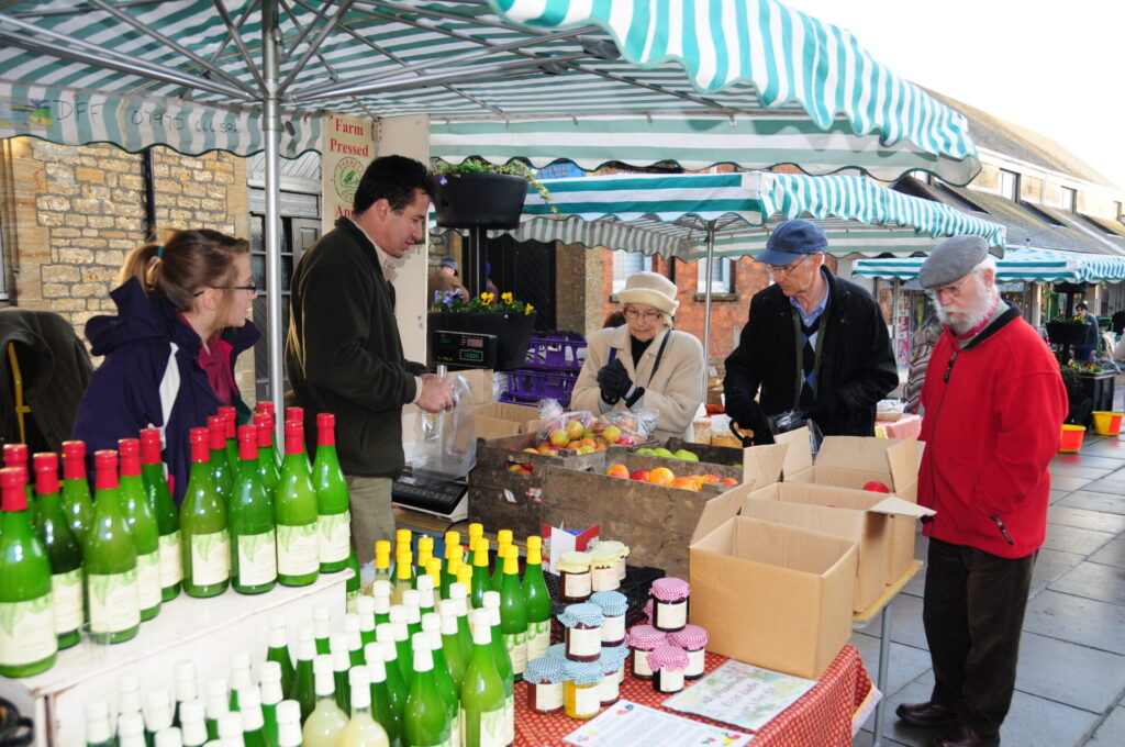 Market Stall with bottles. A man and women selling. Two men and a woman customers